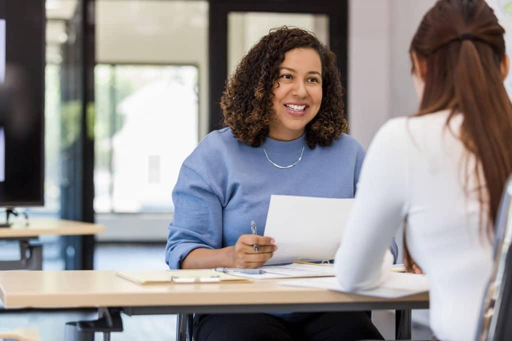 Two women sitting at a desk talking to each other.